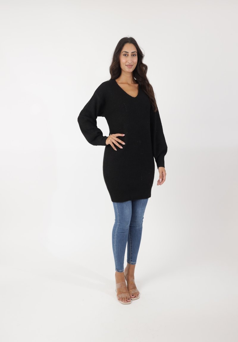Robe pull noire en maille avec un décolleté en V, des manches longues et une taille cintrée, associée à un jean skinny bleu clair et des sandales transparentes.