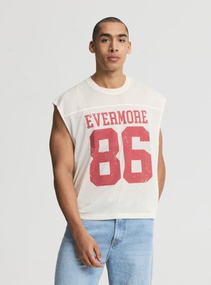 Uomo che indossa una maglietta bianca senza maniche in mesh con scritta rossa "EVERMORE 86" e jeans azzurri, in piedi davanti a uno sfondo semplice.