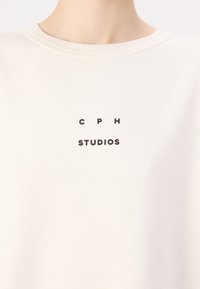 Fehér pulóver kerek nyakkal, amelyen a mellkason középre helyezett fekete nyomtatott szöveg található: "C P H" és "STUDIOS". Puha szövet textúrájú.