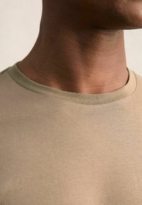 Bēšas krāsas īsām piedurknēm T-krekls ar maigu tekstūru. Apałš kakls izceļas ar tumšāku zaļu akcentu. Audums izskatās gluds ar minimālu šūšanas detaļu.