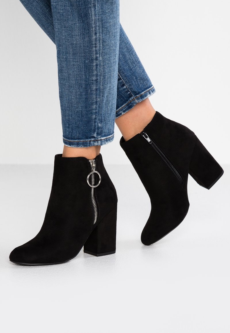 Personne portant un jean bleu et des bottines en daim noir avec talons blocs et fermetures éclair latérales avec languettes rondes.