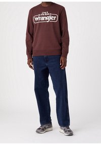 Uomo che indossa un sweatshirt maroon della Wrangler, jeans blu scuro e sneakers grigie della New Balance, in piedi contro uno sfondo bianco semplice.