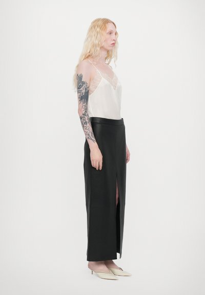 Fustă maxi din piele neagră cu o fanta laterală, asortată cu un top din satin crem cu dantelă. Modelul are părul ondulat, blond și tatuaje.