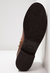 Semelle de chaussure marron avec une bande de roulement en caoutchouc noir texturé et une inscription taille 37, inclinée contre une surface blanche et un mur.