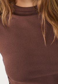 Ruskea, muotoiltu toppi, joka on valmistettu pehmeästä kankaa. Siinä on korkea kaula ja sileä pinta. Ei näkyviä kuvioita tai varusteita.