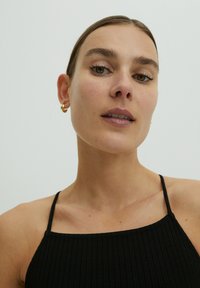 Schwarzes, geripptes Trägershirt mit dünnen Trägern. Das Model hat einen hellen Teint und trägt kleine goldene Creolen. Der Hintergrund ist rein weiß.