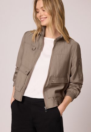 Femme souriante portant une veste légère marron clair zippée avec des poches et une chemise blanche, mains dans les poches d'un pantalon noir, regardant vers le bas sur le côté.