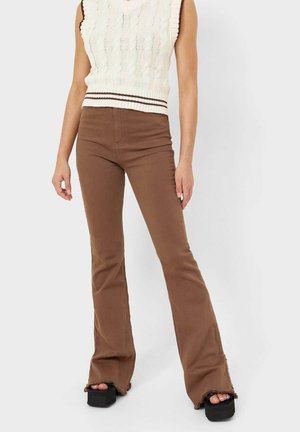 Osoba ubrana w brązowe spodnie dzwony z postrzępionymi brzegami, kremowy bezrękawnik z dzianiny warkoczowej oraz czarne sandały na platformie, stojąca na jednolitym tle.