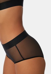 Close-up van een persoon die zwarte transparante hoge taille onderbroek en bijpassende sportbeha draagt, tegen een effen achtergrond.