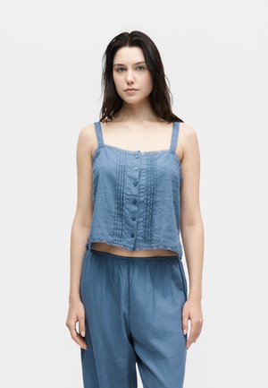 Pikkade tumedate juustega naine kannab sinist varrukateta nööbitavat lühikest pluusi ja lahtisi siniseid pükse, seistes ühevärvilise heleda tausta ees.