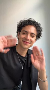 Jeune personne aux cheveux bouclés souriant et saluant des deux mains, portant un t-shirt noir et des bijoux en argent, sur un fond clair uni.