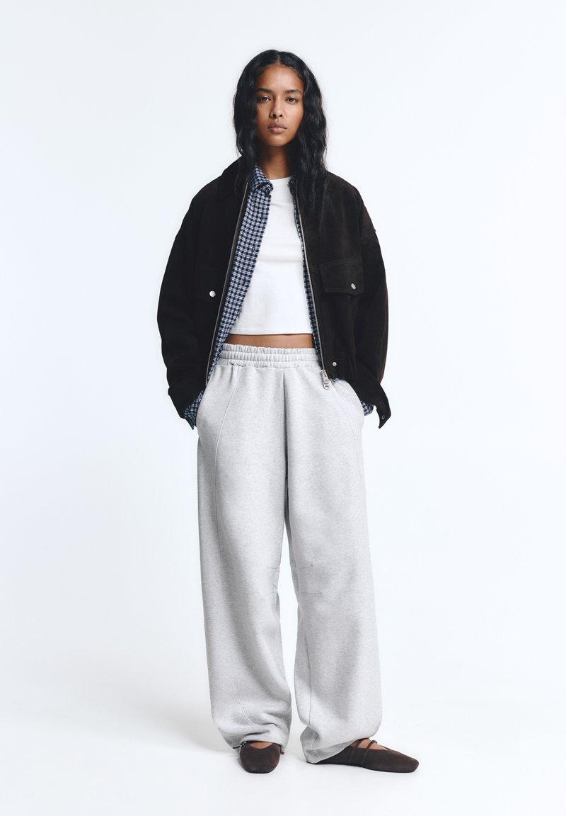 Vrouw die met handen in haar zakken staat, draagt een zwarte jas, geruite blouse, witte crop top, grijze sweatpants en donkere ballerina's op een witte achtergrond.