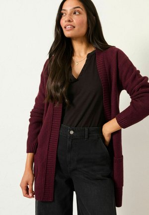 Strickjacke in Burgunderrot mit gerippter Textur, offener Vorderseite und Seitentaschen, getragen über einem schwarzen Henley-Shirt und hoch geschnittenen schwarzen Hosen.