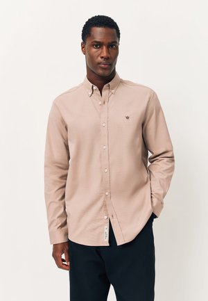 Homme portant une chemise beige à boutons avec un petit logo de couronne sur la poitrine gauche, associée à un pantalon foncé, debout devant un fond uni.