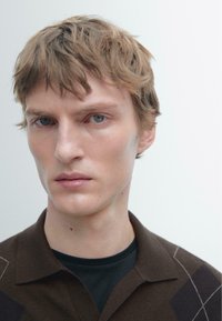 Polo in maglia marrone con colletto, caratterizzata da linee diagonali color crema. Capelli corti e scompigliati, sfondo neutro.