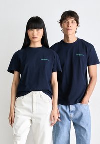 Navy blue cotton t-shirts with "tommy jeans" logo in green, paired with white and light blue denim jeans, displayed on two models.