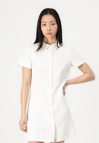 Rochie albă cu mâneci scurte, cu guler, cu nasturi pe față și buzunare laterale; material neted cu o formă ușor structurată, dreaptă.