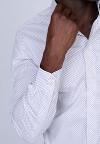 Chemise blanche à boutons avec un petit motif géométrique multicolore. Manches longues avec poignets boutonnés. Tissu au toucher lisse.