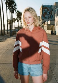 Jonge vrouw met blond haar, een roestkleurig jack met witte strepen en een spijkershorts, staand op een met palmbomen omzoomd pad in zonlicht.