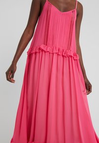 Femme portant une robe sans manches rose vif avec un panneau avant plissé, des insertions en dentelle et une finition à volants à la taille et à l'ourlet sur un fond gris.