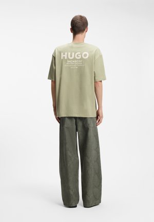 Hombre vestido con camiseta extragrande de color verde claro de la marca HUGO y pantalones de pierna ancha con estampado de serpiente en verde oscuro, de pie de espaldas.