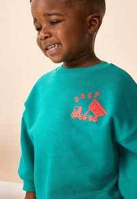 Roheline pusa, millel on tikitud oranž raudtee- ja kallurauto motiiv ning sõna "BEEP" selle kohal. Pehme tekstuur ja lahtine lõige.