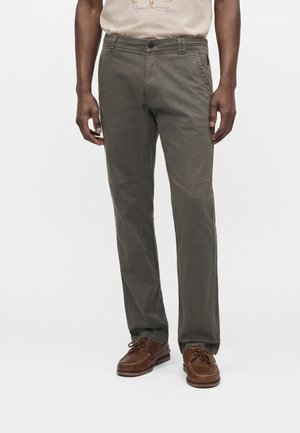 Homme portant un pantalon droit vert olive, des mocassins en cuir marron et un t-shirt beige, debout devant un fond blanc.