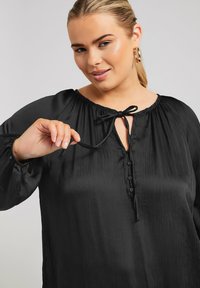 Svart blus tillverkad av slät tyg med en rynkad halsringning, knäppning framtill och en knytdetalj. Långa ärmar och avslappnad passform.