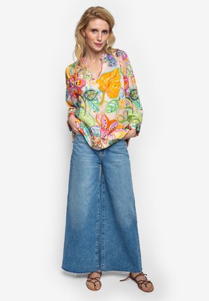 Frau trägt eine bunte, geblümte Bluse, einen weit geschnittenen blauen Jeansrock und braune Sandalen mit Riemchen, steht mit den Händen in den Taschen.