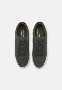 Zapatillas de ante verde con puntera redonda, diseño con cordones y suela de caucho blanca contrastante. Forro interior en verde oscuro. Branding minimalista visible.