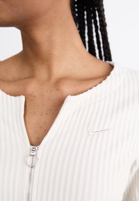White ribbed zip-up top with a round neckline, featuring a small embroidered Nike logo on the left chest and a silver zipper pull.