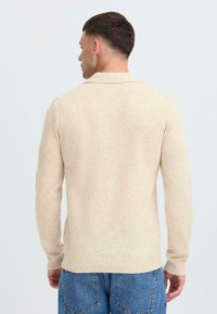 Maglione beige a maglia con maniche lunghe e colletto. Texture morbida, design aderente e polsini a coste. Indossato con jeans in denim blu.