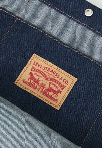 Levi Strauss & Co. etiķete brūnā ādā uz tumšajiem džinsiem, kurā ir sarkani drukāti zīmējumi ar zirgiem un tekstu. Ir redzama kontrastējoša šūšana.