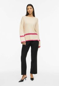 Femme debout portant un pull beige à rayures roses, un pantalon noir droit et des chaussures à talons aiguilles noires sur un fond blanc.