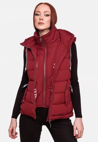 Crvena puffer prsluk s visokim ovratnikom, zatvaranjem na zip i uvučenim detaljima. Ima quilted teksturu i bočne džepove. Nositi preko crne majice s dugim rukavima.