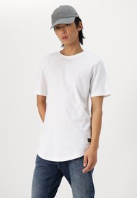 T-shirt blanc à manches courtes avec un col rond, associé à un jeans droit bleu foncé. Le look est complété par une casquette de baseball grise.