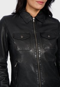 Black leather jacket with a collared neckline, front zipper, two chest pockets with flaps, and two side pockets. Smooth texture and fitted design.
