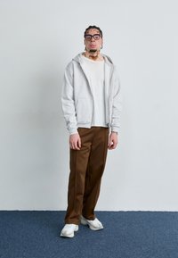 Grå zip-up hoodie over en hvid t-shirt, parret med brune bukser og hvide sneakers. Accessories inkluderer briller og et armbånd.