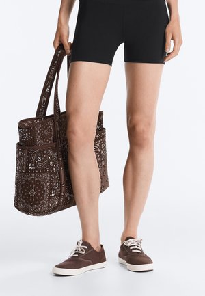 Persoon draagt zwarte korte broek en bruine sneakers en houdt een grote bruine tas met patroon naast zich vast.