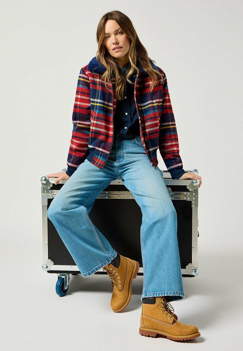 Giacca di flanella a quadri rossa, blu e gialla, con colletto blu, abbinata a jeans chiari a vita alta con fondo svasato e stivali color sabbia.