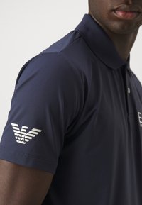 Navy blue polo shirt featuring a collar and short sleeves, with a textured fabric and a white logo on the left sleeve.
