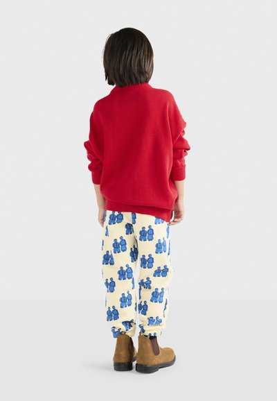 Rød strikket sweater, cremefarvede bukser med blå figurmønstre, og brune ankelstøvler. Synlig manchet detalje på sweatre og elastiske manchetter på bukserne.