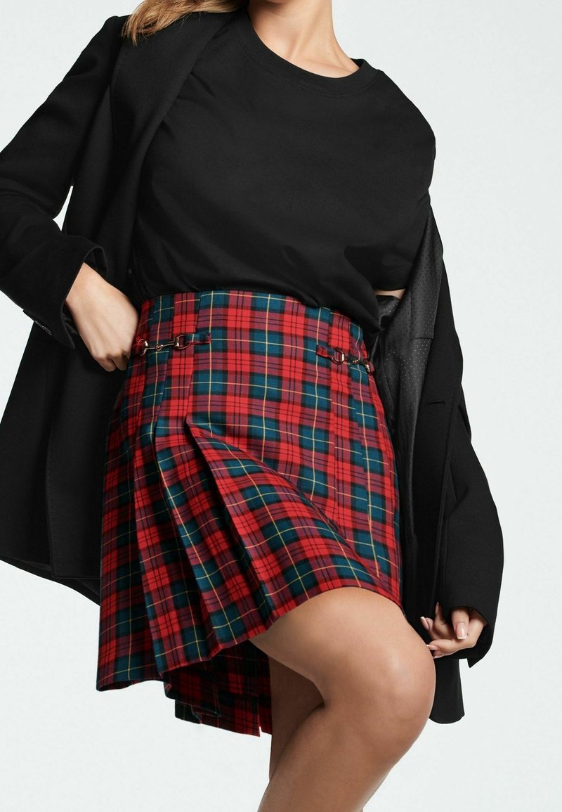 Femme portant un haut noir et un blazer avec une jupe plissée à carreaux rouges et bleus avec des boucles latérales, posant avec une main sur la hanche.