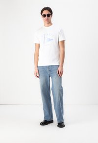 White t-shirt with blue "HACKETT" logo and graphic, paired with light blue straight-leg jeans and black shoes. Black sunglasses worn.