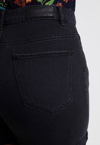 Pantalones cortos de mezclilla negra con un bolsillo trasero, costuras visibles y una etiqueta de cuero en la cintura. La tela tiene una textura suave.