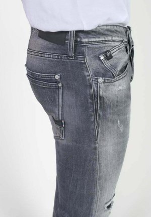 Jeans en denim gris avec une coupe slim, des détails effilochés, plusieurs poches et une texture lisse. Marque emblématique sur la ceinture.