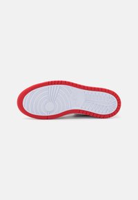 Semelle en caoutchouc blanche et rouge d'une chaussure Nike montrant des motifs de crampons circulaires et linéaires, vue par en dessous sur un fond uni.