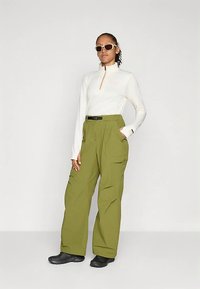 Top bianco a maniche lunghe con cerniera e logo, abbinato a pantaloni cargo larghi verde oliva e stivali neri; accessoriato con occhiali da sole chiari.