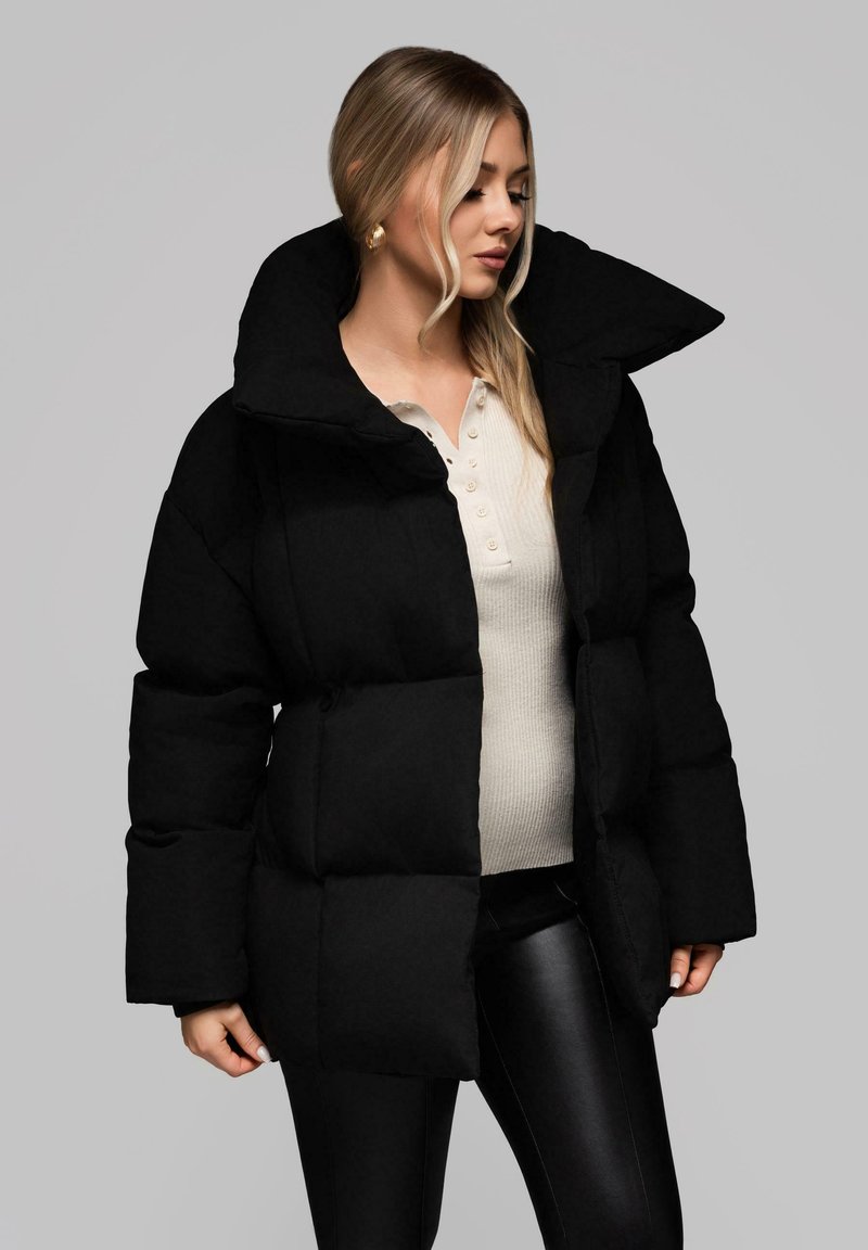 Černá vycpaná péřová bunda s širokým límcem, oversized siluetou a texturovaným materiálem. Nositelná přes světle béžový top a černé kalhoty.