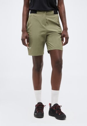 Άτομο που φοράει χακί σορτς, μαύρο πουκάμισο, λευκές κάλτσες και μαύρα παπούτσια πεζοπορίας με κόκκινα κορδόνια, στέκεται μπροστά σε απλό φόντο.